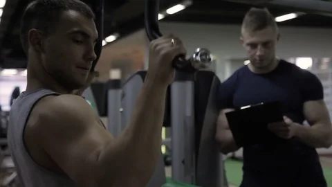 Young bodybuilder is doing wide grip pulldowns exercise with coach in gym. Stock Footage 85227843