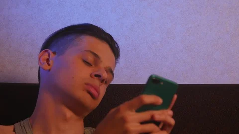 Young bored guy sitting on the sofa in the room and using mobile phone Video stock 106371325