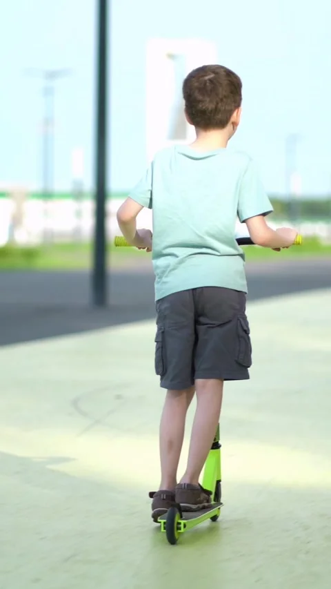 Young boy from behind rides a bright green scooter on a sunny paved path outdoor Stock Footage 341530545