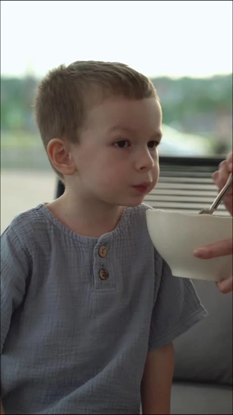 Young boy being fed soup by his mother with a spoon on the veranda Vídeo Stock 336868785