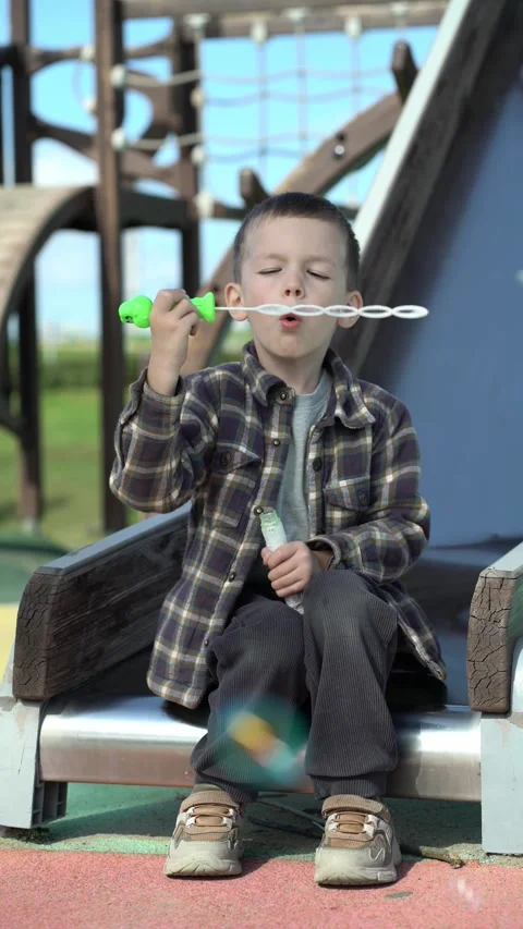 Young boy blowing soap bubbles at playground. Cute child sits outdoors creating Stock Footage 345528001