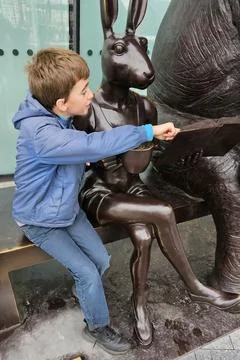 Young boy in a blue jacket sits beside a bronze rabbit statue, pointing at an Stock Photos