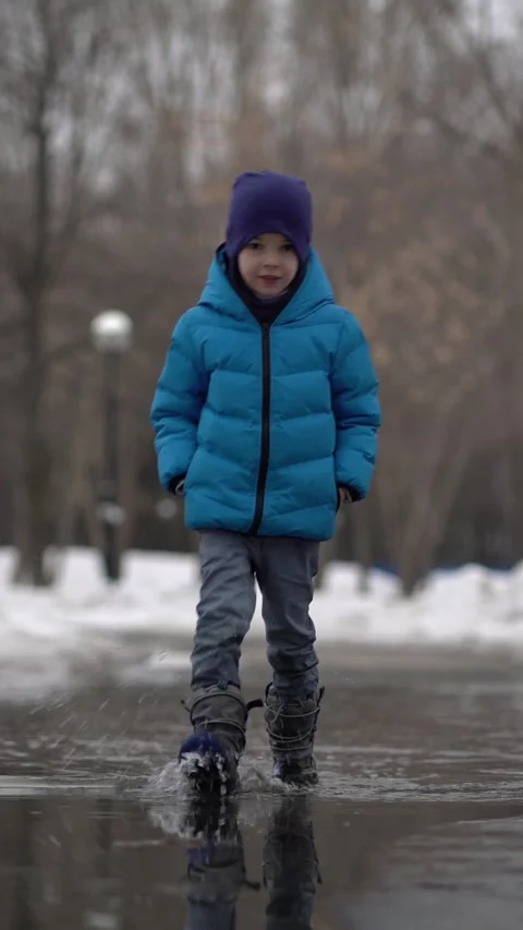 Young boy in blue puffer jacket and beanie walks through a puddle on a snowy Stock Footage 341530604