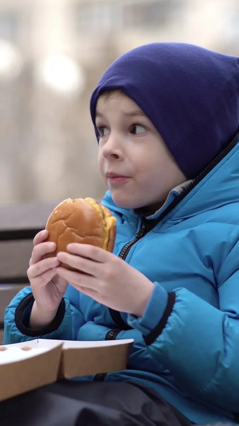Young boy in blue winter jacket and beanie happily eating a cheeseburger Vídeo Stock 348790147