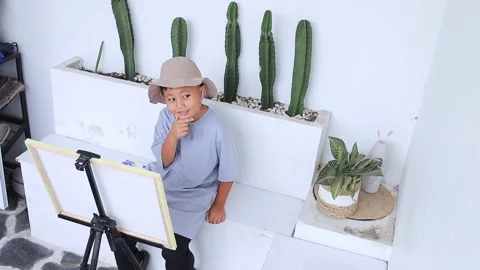 Young boy in bucket hat sitting next to cacti while looking thoughtfully at his 스톡 동영상 324947959