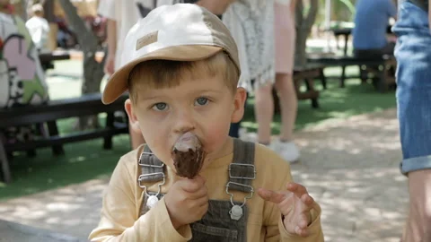 Young boy in cap enjoys chocolate ice cream outdoors, getting messy while eating Stock Footage 302542945