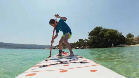 Young boy confidently paddling on a stand-up paddleboard in shallow turquoise Stock Footage 313808451