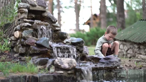 Young boy crouches by a stone waterfall in a pine forest, Stock Footage 340933216