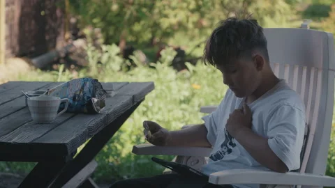 Young boy eating potato chips while using computer tablet at garden picnic table Stock Footage 337767895