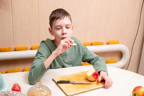 A young boy eating a slice of a red apple at a table with a wooden cutting .. Stock Photos