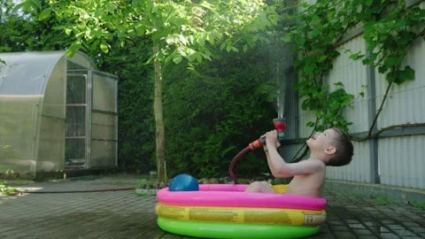 Young boy enjoying summer fun in inflatable pool with water spray and greenery Stock Footage 313333521