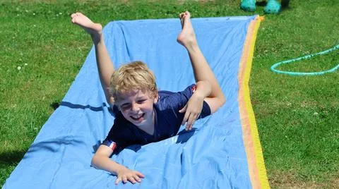 Young boy enjoys sliding down a wet blue slip-and-slide on a grassy lawn Stock Photos