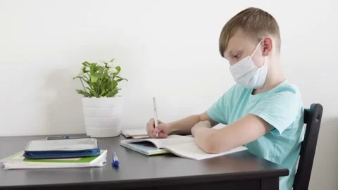 A young boy in a face mask does homework for school at a table at home 스톡 동영상 138164356
