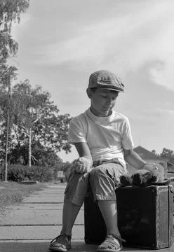 Young boy in flat cap sits on vintage suitcase with teddy bear, looking tho.. Stock Photos