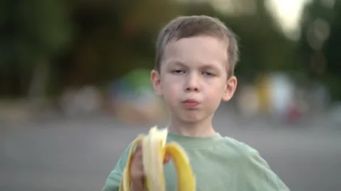 Young boy in green shirt eats a peeled banana outdoors on a sunny street Video stock 340670439