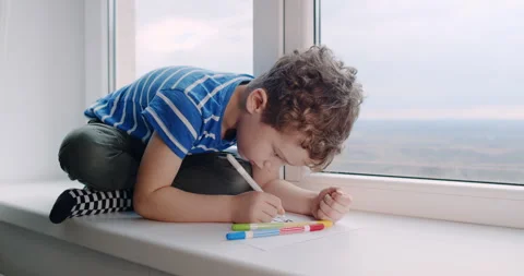 A young boy is happily sitting on a window sill drawing with colorful markers Stock Footage 307297539