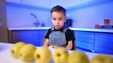 Young boy holding bitten yellow apple at counter in blue illuminated kitchen Stock Footage 342161061