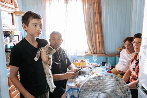 Young boy holding a cat stands in a brightly lit kitchen with family member.. Stock Photos