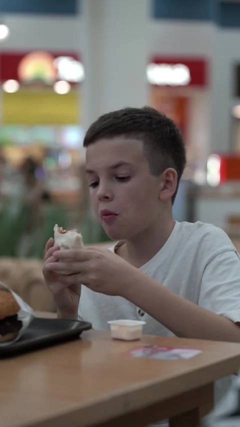 Young boy holding wrap and looking aside while seated at food court Video stock 343455628