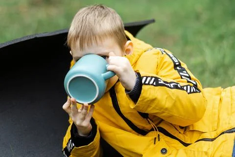 A young boy in a jacket drinks from a cup in the green forest at a picnic. Stock Photos