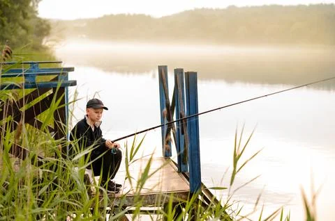 Young boy learning to fish alone on a wooden pier by a misty lake Stock Photos