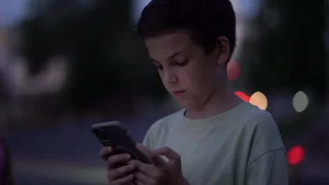 Young boy in light green shirt focused on smartphone outdoors at night  Stock Footage 340670672
