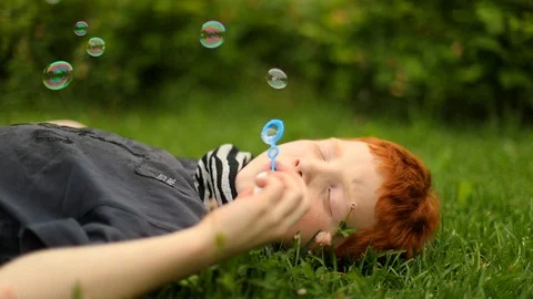 Young boy lying on grass in the park and blowing bubbles, slow motion Stock Footage 98027329