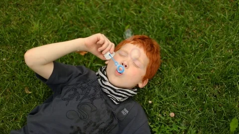 Young boy lying on grass in the park and blowing bubbles, slow motion Vídeos de archivo 98027365
