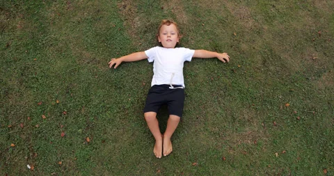 Young boy lying on grass, rubs bare feet against the short lawn, high angle shot Vidéo 322045782