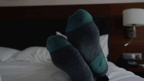 The young boy is lying on a white bed in socks and wireless headphones and looks 動画素材 114798285