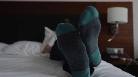 The young boy is lying on a white bed in socks and wireless headphones and looks 動画素材 115069313