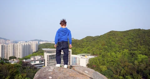 Young boy make step and standing on a rock overlooking a city and green hills Stock Footage 322098902