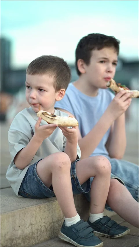 Young boy with messy face eats pizza slice on outdoor steps while older brothe Video stock 337678065