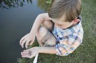 A Young Boy Outdoors Sitting On A Riverbank With A Small Fish In The Palm Of Stock Photos