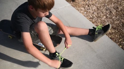 Young boy at park sitting on a skateboard tying his shoe Stock Footage 94930341