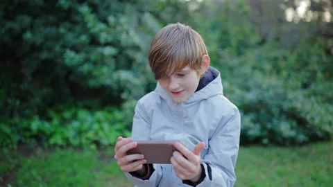 Young Boy Playing Mobile Game on Smartphone in Green Park at Sunset. Vidéo 338140545