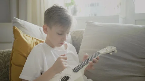 Young boy playing white ukulele on a sofa at his home Stock-Footage 128362780