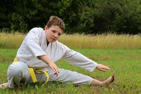 Young boy practices karate stretching in a grassy field during daylight hours 库存照片