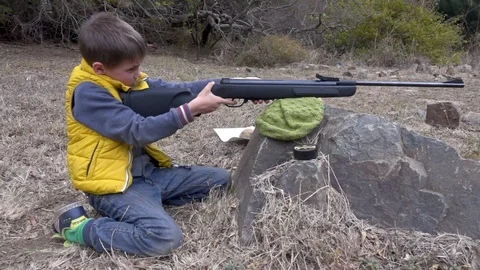 Young boy pulls the trigger on small air rifle leaning against a rock in slow Vídeo Stock 104064324