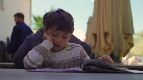 Young boy reading book at outdoor cafe table with thoughtful expression and.. Stock Footage 335681376