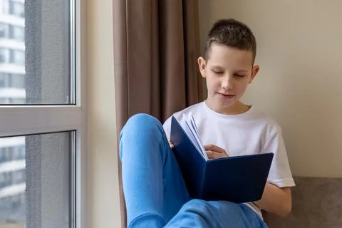 Young Boy Reading a Book by Window. A young boy in a white shirt and blue pants Stock Photos