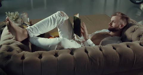 A young boy reads a book lying in the apartment on the couch. A man is learning. Stock Footage 145022093