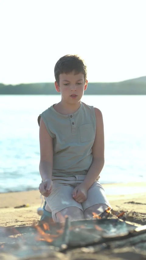 Young boy roasting marshmallow over campfire on sandy beach by lake at sunset. Stock Footage 341989594