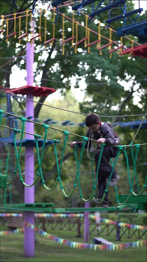 A young boy in a safety harness carefully walks across a hanging green rope Stock Footage 345285278