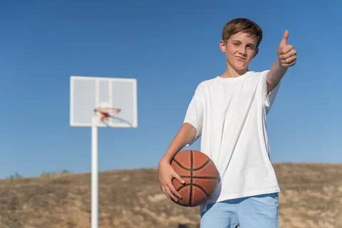 Young boy showing thumb up while posing with basketball ball at an outdoor court Stock-Fotos