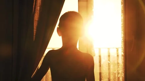 Young boy silhouette observes warm sunbeams from cozy balcony in morning Stock Footage 327087149