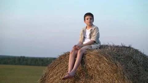 Young boy sits barefoot atop a large hay bale in a quiet rural field at dusk Vídeo Stock 342966785