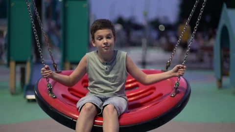 Young boy sits on a bright red circular swing in a playground at dusk Stock Footage 341410035