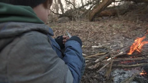 A young boy sits by a campfire in the forest, carving wood with a knife. This Stock Footage 290874818