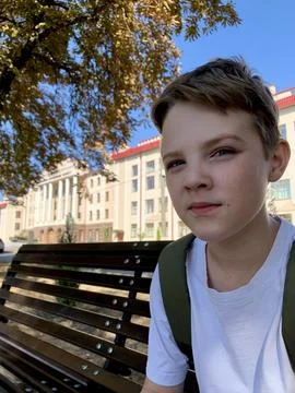 A young boy sits casually on a bench, wearing a white shirt and backpack, in Stock Photos